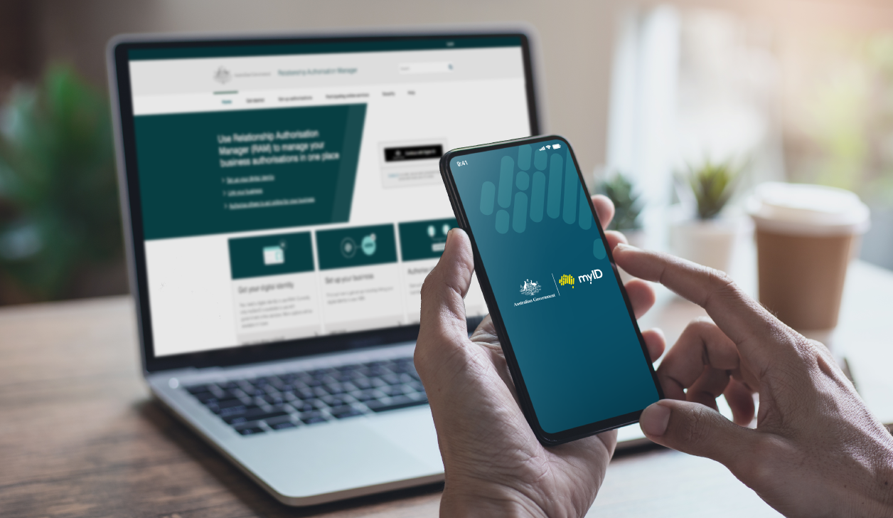 Hand holding phone with the myID app open in the foreground, laptop in the background showing the Relationship Authorisation Manager information site.